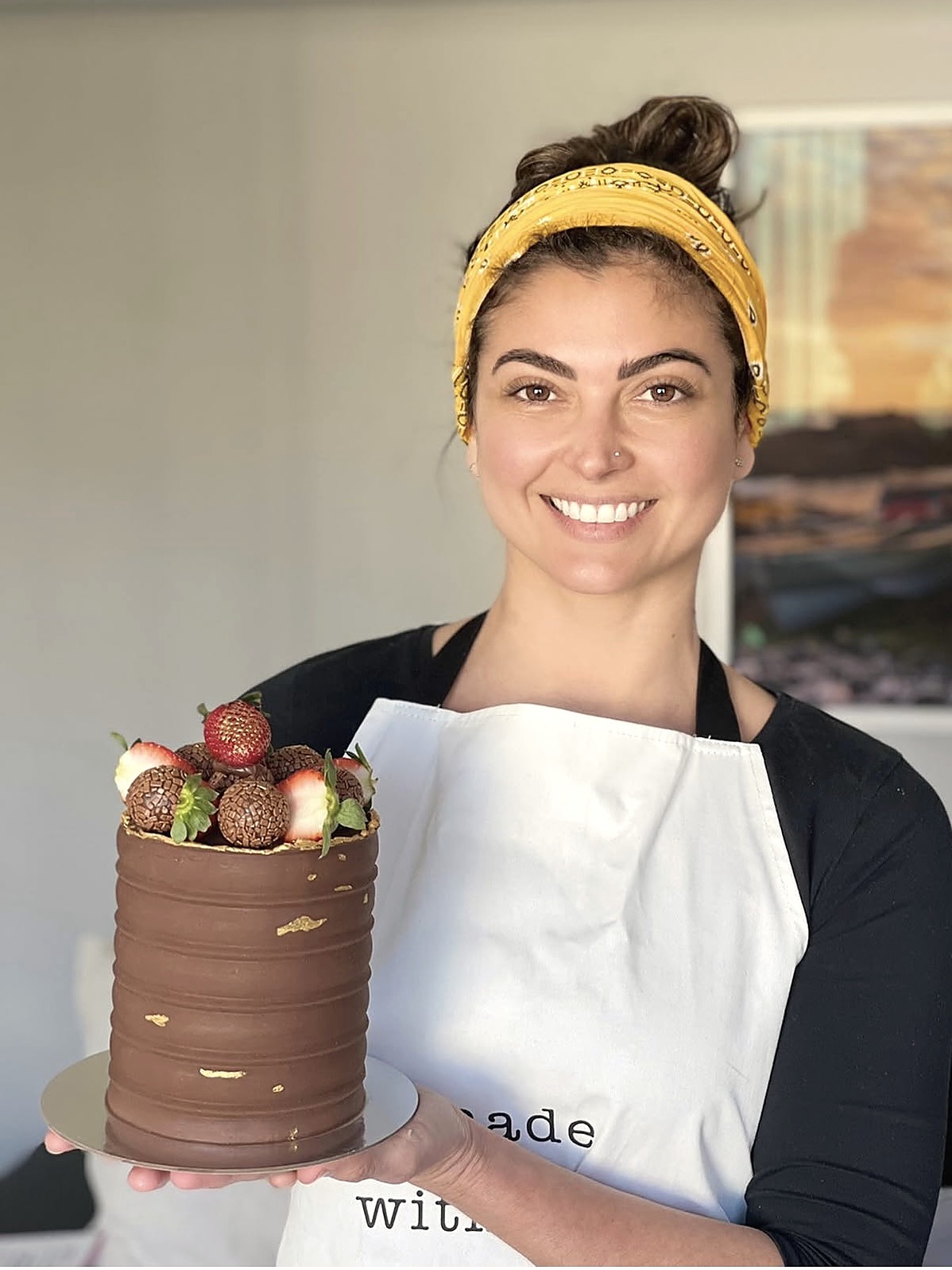 Noemi, the baker behind Cakes Delight, smiling and holding a chocolate cake topped with strawberries